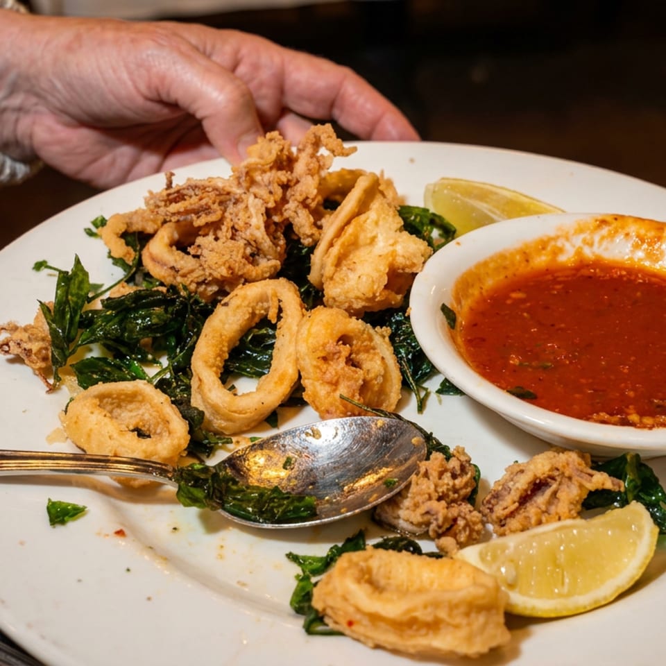 A hand serves a plate of crispy fried calamari with lemon wedges and a side of red sauce.
