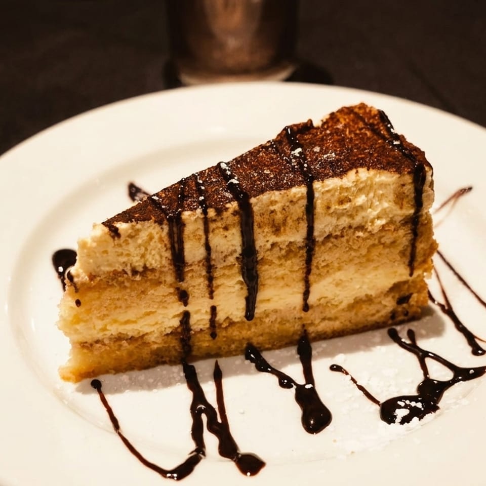 A slice of tiramisu with chocolate drizzle and powdered sugar on a white plate.