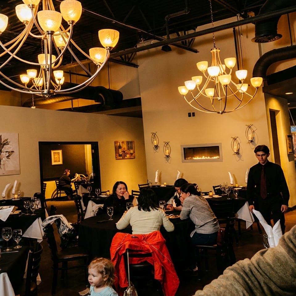 Restaurant dining room with patrons seated at tables, waiters, and elegant chandeliers overhead.