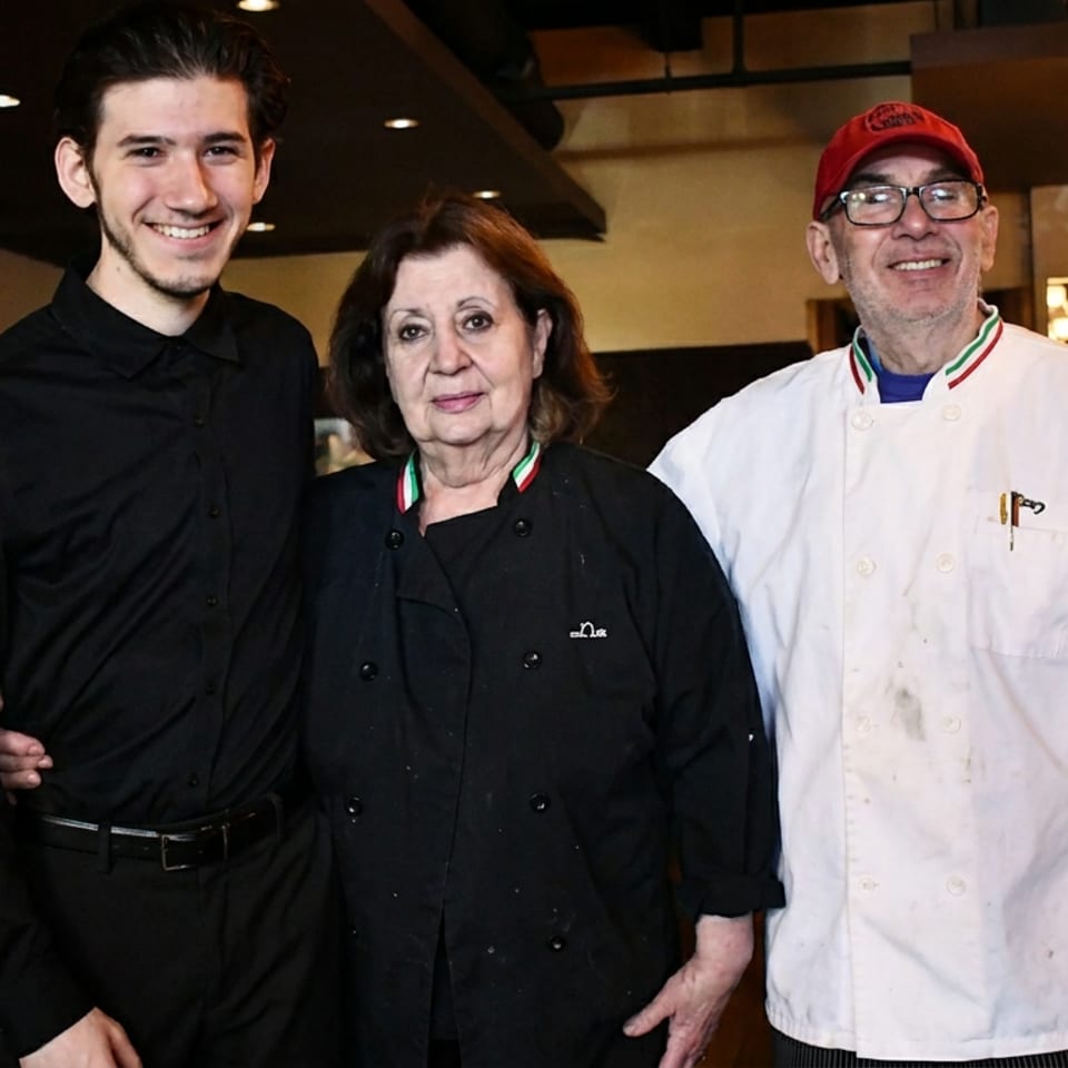Three smiling restaurant staff, two chefs and a server, standing in a dining room.