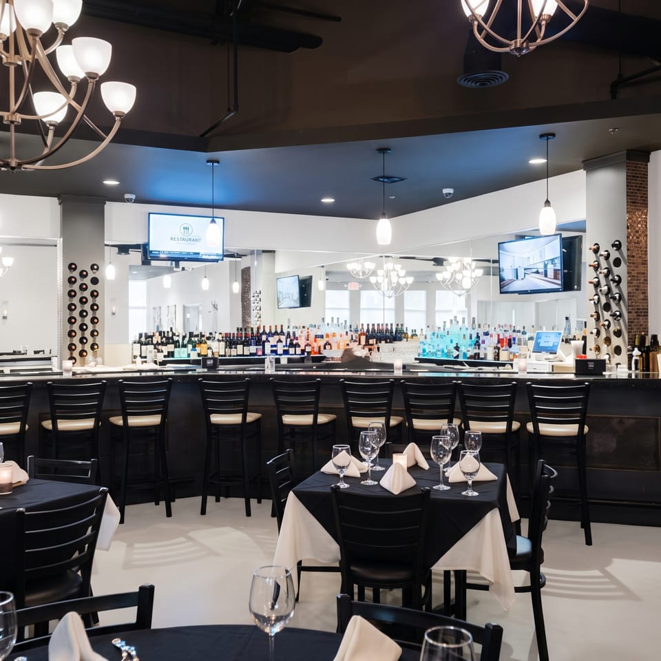Elegant restaurant dining room with black tablecloths, set tables, chandelier lighting, a stocked bar, and wine displays.